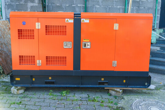Outdoor industrial generator with orange casing on concrete blocks near brick wall