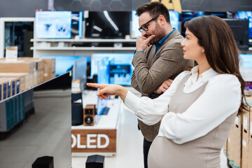 Fototapeta premium A smiling young couple shops for home entertainment, looking at a large flat screen television in a modern tech retail store department.