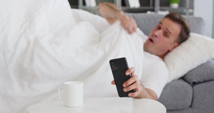 Morning man silences phone alarm, tired wakeup. Sleepy adult in bed on a sofa stops the ringtone, hits snooze, and delays getting up, risking being late. Clear concept: waking late with phone.