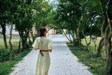 Obraz premium Woman in a pale dress walks along a gravel path under lush trees in a peaceful park, conveying calm, exploration and a timeless outdoor moment