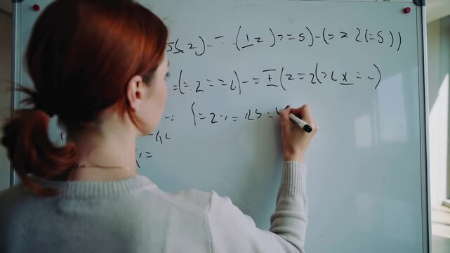 STEM professional solving algebraic problems on large whiteboard. Redhead woman in gray sweater working in bright modern office space.