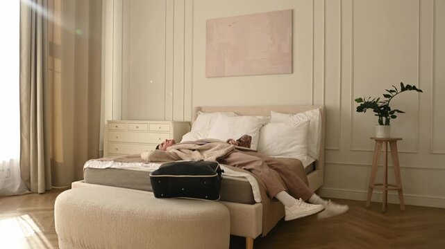Young exhausted woman arriving at her destination, entering a stylish hotel room or apartment bedroom, and immediately falling backwards onto the comfortable bed to rest after a long trip