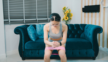 Obraz premium Young woman exercising with resistance band in living room, focused on fitness and strength training, casual home workout with comfortable sofa and bright sunflowers in background