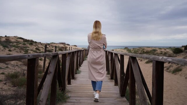 Rear view of blonde woman in trench coat walk along wooden pathway through sand dunes, heading towards sea on cloudy day, embody sense of freedom and solitude