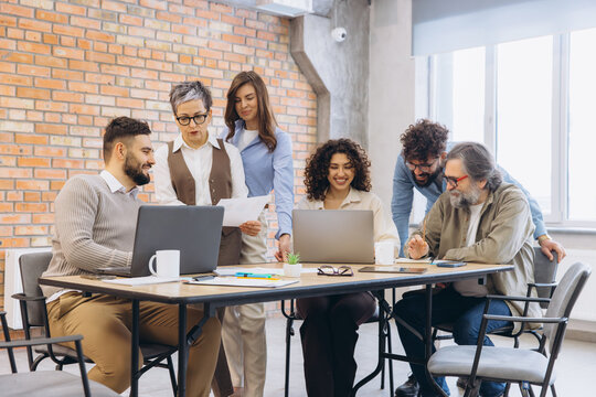 Diverse team collaborating during business meeting at modern office, discussing work together with laptops and papers