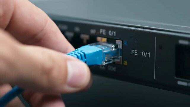 Close up of a hand plugging a blue ethernet cable into a network switch port.
