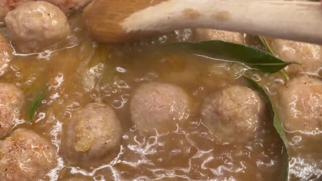 Close-up of meatballs simmering in rich brown curry sauce with kaffir lime leaves, stirred with wooden spoon in pot.