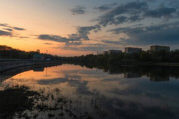 Obraz premium A peaceful evening on Minsk lakeside. Sunset sky, painted in hues of orange and purple, is mirrored perfectly in the calm water, with the illuminated city skyline creating a modern silhouette.