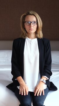 Young businesswoman with glasses experiencing a panic attack, breathing heavily while sitting on a bed in a hotel room after work, feeling stressed, anxious, and overwhelmed by her problems