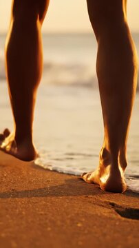 Walking on sandy beach at sunset. Barefoot legs step gently on shoreline. Ocean waves touch feet, creating a serene, peaceful atmosphere. Reflection of golden light enhances natural beauty.