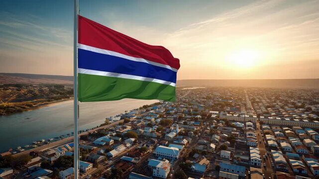 The Gambian flag flies high over a bustling West African city as the sun sets. The scene shows a vibrant community near a river and numerous boats nearby.