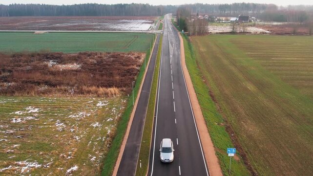 A van and car pass each other on a well-paved rural highway flanked by winter fields with patches of snow. Aerial perspective.