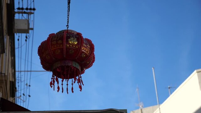 Red Chinese lantern with gold details and tassels hanging outdoors against a blue
sky, urban architecture background. This description is formulated as someone would
search for it online.

