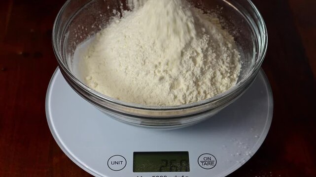 a steady stream of white flour being poured into a clear glass bowl resting on a digital kitchen scale.