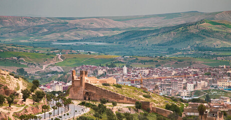 Obraz premium Historic walls of Fez overlooking the medina, ancient architecture and lively city, green hills in the background, contrast between heritage and natural landscape.