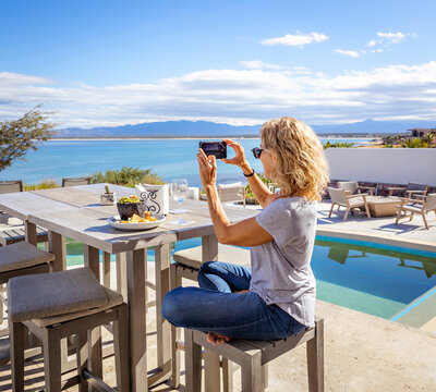 Blonde woman taking a cell phone photo of the ocean from her outdoor table by a pool. 