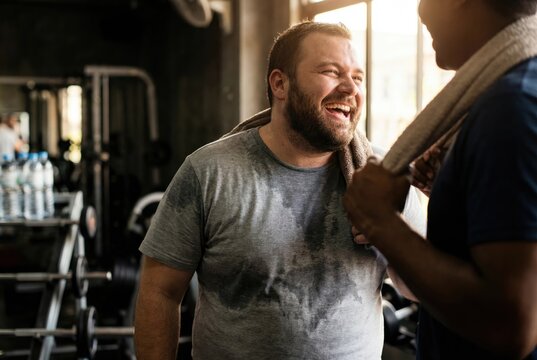 Happy overweight man with a beard laughing with his personal trainer or friend after a rewarding workout session in the gym.
