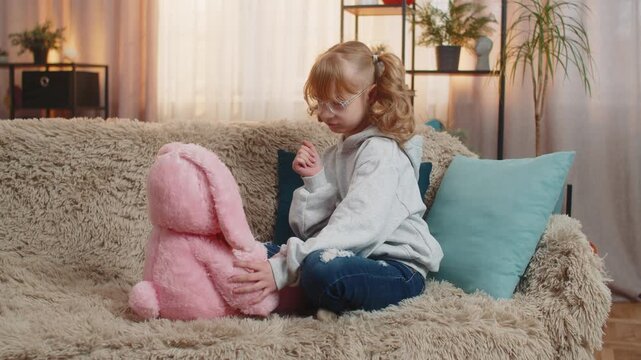 Young teenager girl plays with soft rabbit toy at home, chatting in a playful manner. Blonde child kid on sofa continues warm imaginary talk with gentle affection, smiling sweetly at plush friend.