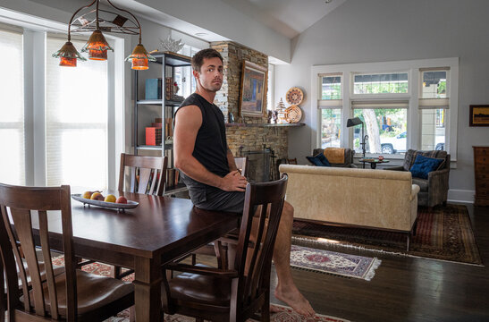 Young strong athletic man with tattoos at a dining room table inside a house. 