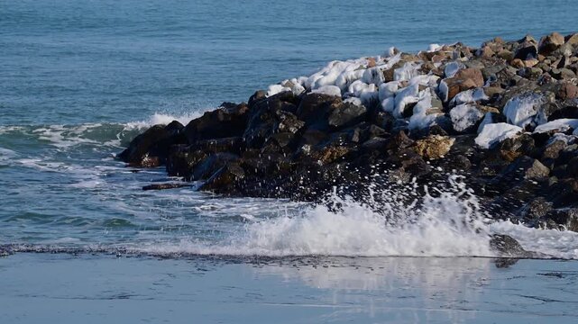 turquoise ocean waves crashing against a rocky breakwater partially covered in patches of white snow.