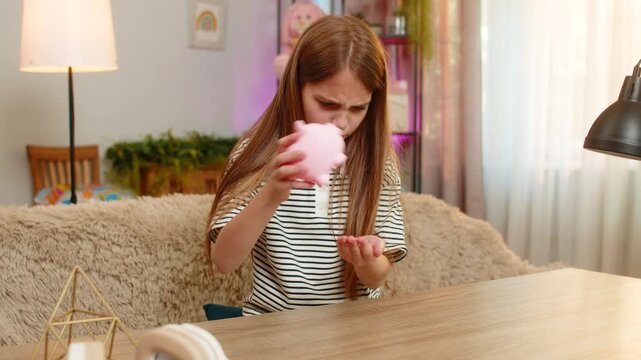 Young child girl sitting at table on home sofa looks at empty piggy bank and shakes head in frustration and sadness. School kid shows disappointment after realizing no money is left for desired toy.