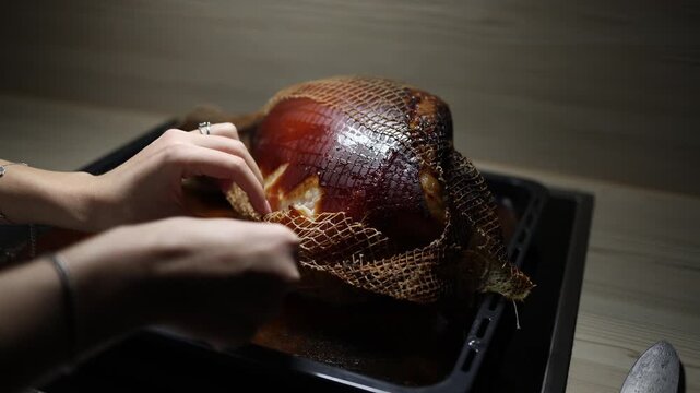 Woman hands gently remove cooking net from roasted pork knuckle on baking tray cozy kitchen interior low light home dinner preparation slow food ritual warmth and care