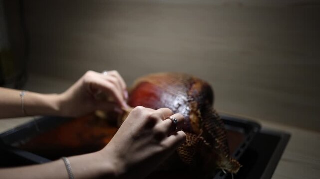 Female hands remove roasting net from baked pork knuckle on tray warm home kitchen soft dim light slow cooking process comfort food tradition evening meal atmosphere