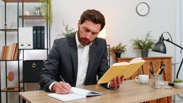 Middle-aged businessman at home office reads leadership book finds quote smiles and notes write idea in notebook. Freelancer guy at table writes plan sets goals for growth, feels motivated by insight