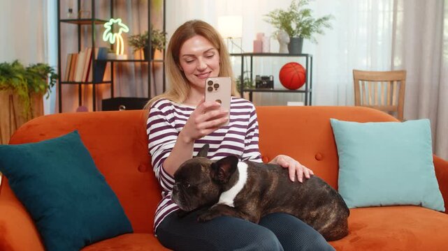 Middle-aged woman holds French bulldog at home scrolls social media feed on smartphone feels calm. Girl dog on sofa watches screen reads posts likes comments gives care and support morning routine joy