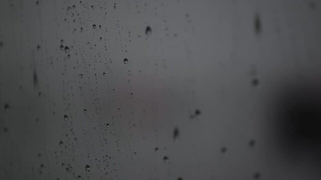 Rain falls on a window, creating drops and streaks during a cloudy day in a quiet city
