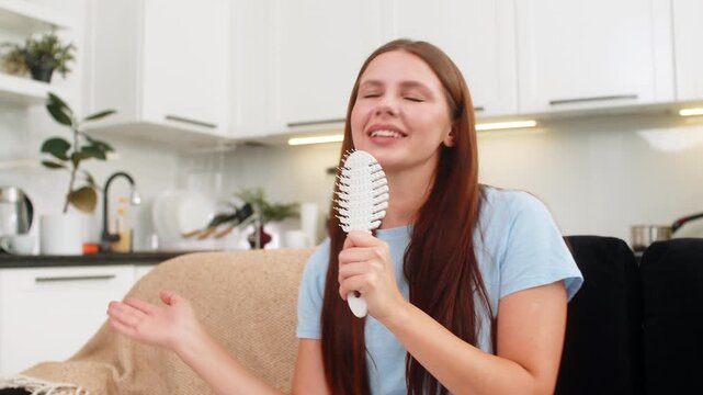 Young woman at home performs pop idol fantasy sings into comb as mic and dances to hit track with fun. Caucasian girl on sofa laughs sways claps enjoys music play and keeps bright happy mood and joy.