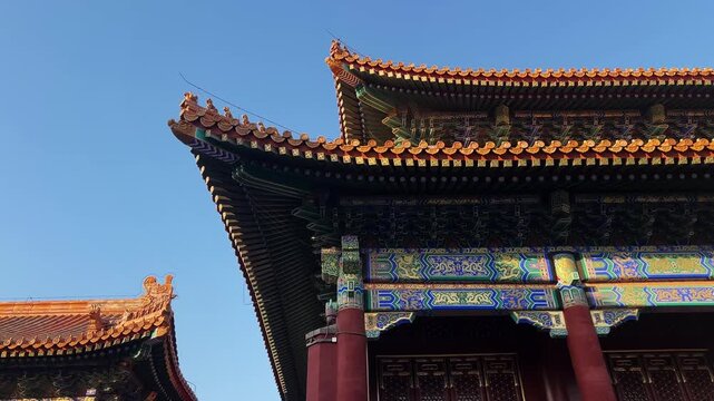 Low angle shot shows ornate roof corner and painted beams of Forbidden City palace building in Beijing, with golden roof tiles and intricate dougong brackets against clear blue sky.