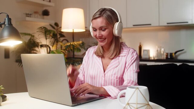 Adult woman at night home works in design tool on laptop with headphones moves layers tweaks layout smiles. Caucasian girl at table on sofa at evening nods music applies edits saves version progress.