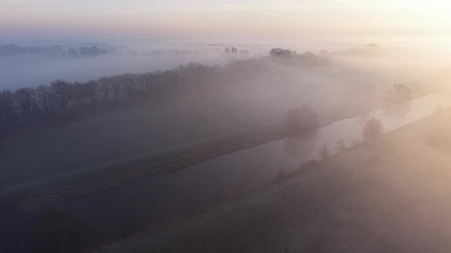 A misty winter landscape with a narrow river reflecting soft light frosty fields and bare trees fading into dense fog creating a calm and dreamy sunrise atmosphere.