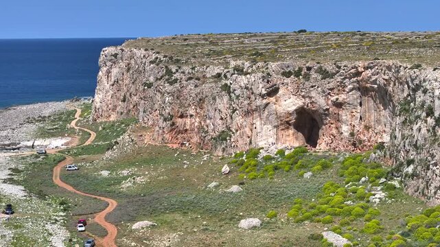 AERIAL: Towering limestone cliff with a large cave and a winding dirt road below. Tiny vehicles are parked at a popular rock climbing destination near San Vito Lo Capo on scenic island of Sicily.