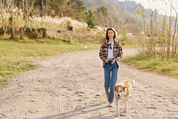 Fototapeta premium Woman walking on a dirt road with a leash dog in a rural setting, casual attire and plaid shirt, jeans, enjoying nature and daylight with a calm countryside mood.