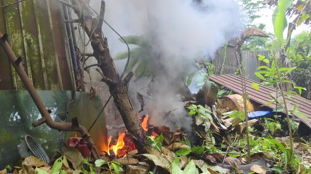 Intense outdoor fire burning a tree trunk amid thick white smoke, surrounded by dry banana leaves, branches, debris, rusty metal sheets, and scattered vegetation in a backyard-like area.