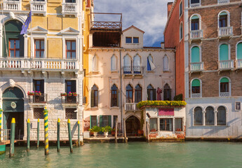 Obraz premium Colorful historic buildings line the edge of a canal in Venice, Italy, with arched windows, balconies, and mooring poles.