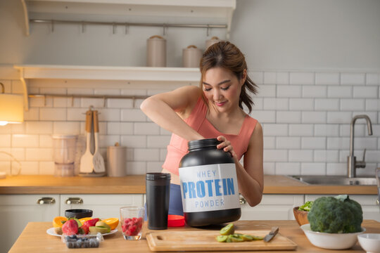Asian woman scooping whey protein powder into shaker bottle and healthy vegetables in kitchen