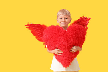 Obraz premium Cute little happy boy dressed as cupid with heart-shaped fluffy pillow on yellow background. Valentine's Day celebration