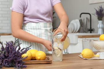 Young woman pouring lemon juice from jug into glass for preparing lavender lemonade at table in...