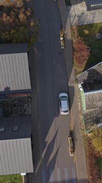 Vertical Screen Aerial Topdown Of Small Town Street With Car And Delivery Activity Near Shopfronts, Long Pedestrian Shadows And Curbside Parking Showing Everyday Commerce And Route Movement