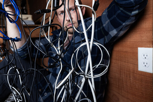 A stressed adult male struggles with a chaotic tangle of various electronic cables, USB cords, and chargers in a dark room. Conceptual image for technology frustration, IT problems, and the need for o