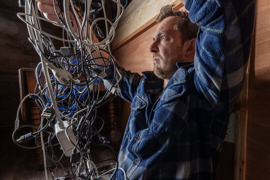 A stressed adult male struggles with a chaotic tangle of various electronic cables, USB cords, and chargers in a dark room. Conceptual image for technology frustration, IT problems, and the need for o