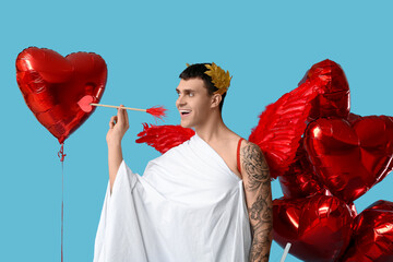 Obraz premium Young man dressed as Cupid with arrow and balloons on blue background. Valentine's Day celebration
