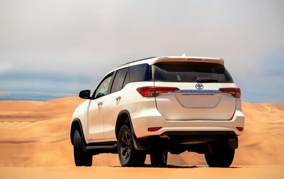 A white Toyota Fortuner parked among golden sand dunes of the Namib Desert, illuminated by bright summer sunlight. A powerful symbol of off-road adventure, freedom, and rugged outdoor elegance. 