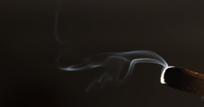Swirling patterns of smoke drift from a glowing palo santo stick, highlighted by angled sunlight in a dark environment. 