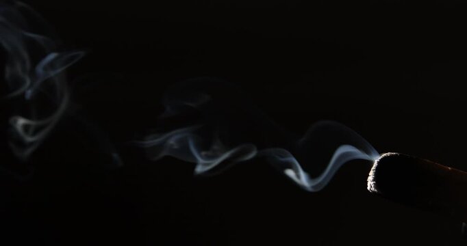 Swirling patterns of smoke drift from a glowing palo santo stick, highlighted by angled sunlight in a dark environment. 