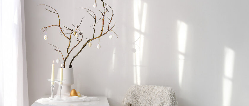 Vase with tree branches, Easter eggs and burning candles on table in light room