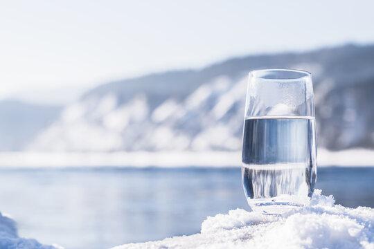 A glass filled with clean natural artesian water sits on snow. In the background, there is a winter landscape with mountains and clear blue skies copy space nature . High quality photo